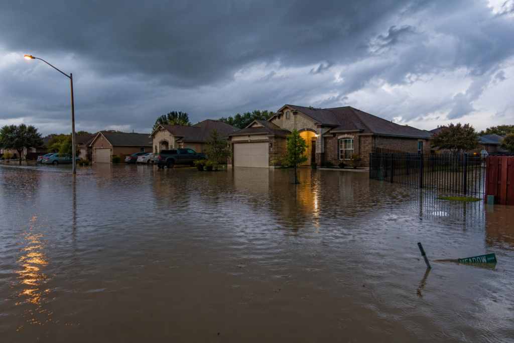 Flooded residential neighborhood in Killeen, Texas after a severe rainstorm, with streets and front yards submerged in water, homes surrounded by floodwater, and dark storm clouds overhead.